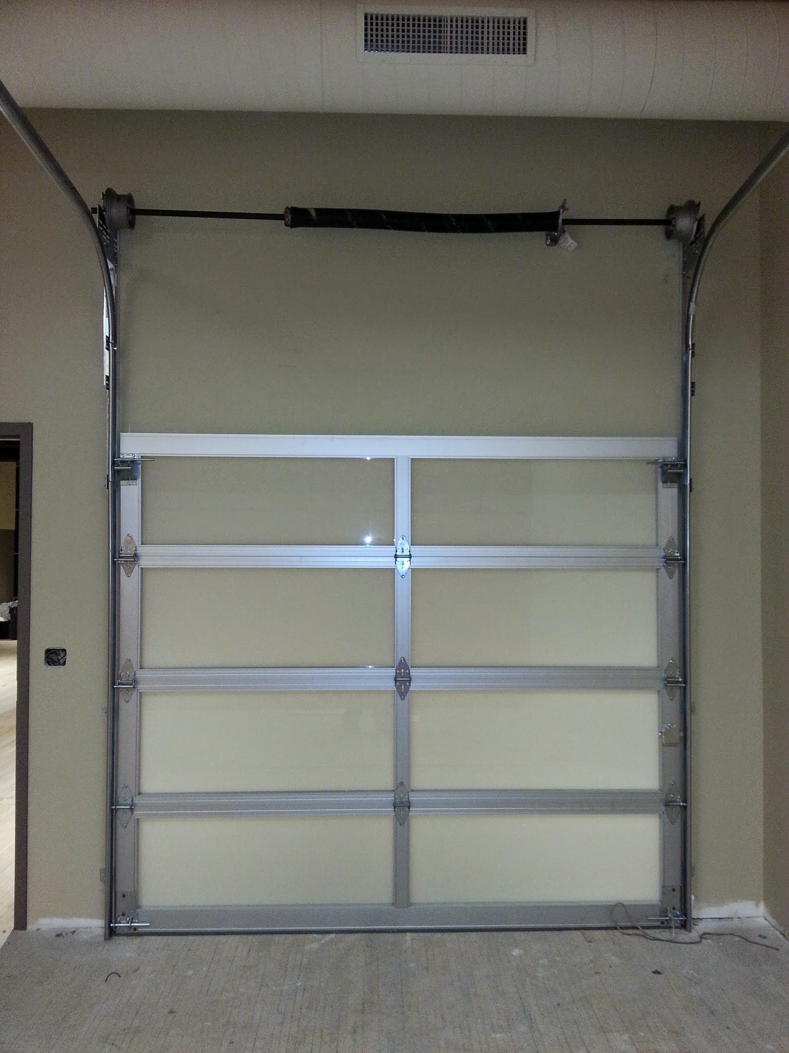 A tall full-view glass garage door with an aluminum frame, seen from inside with its track and opener rail