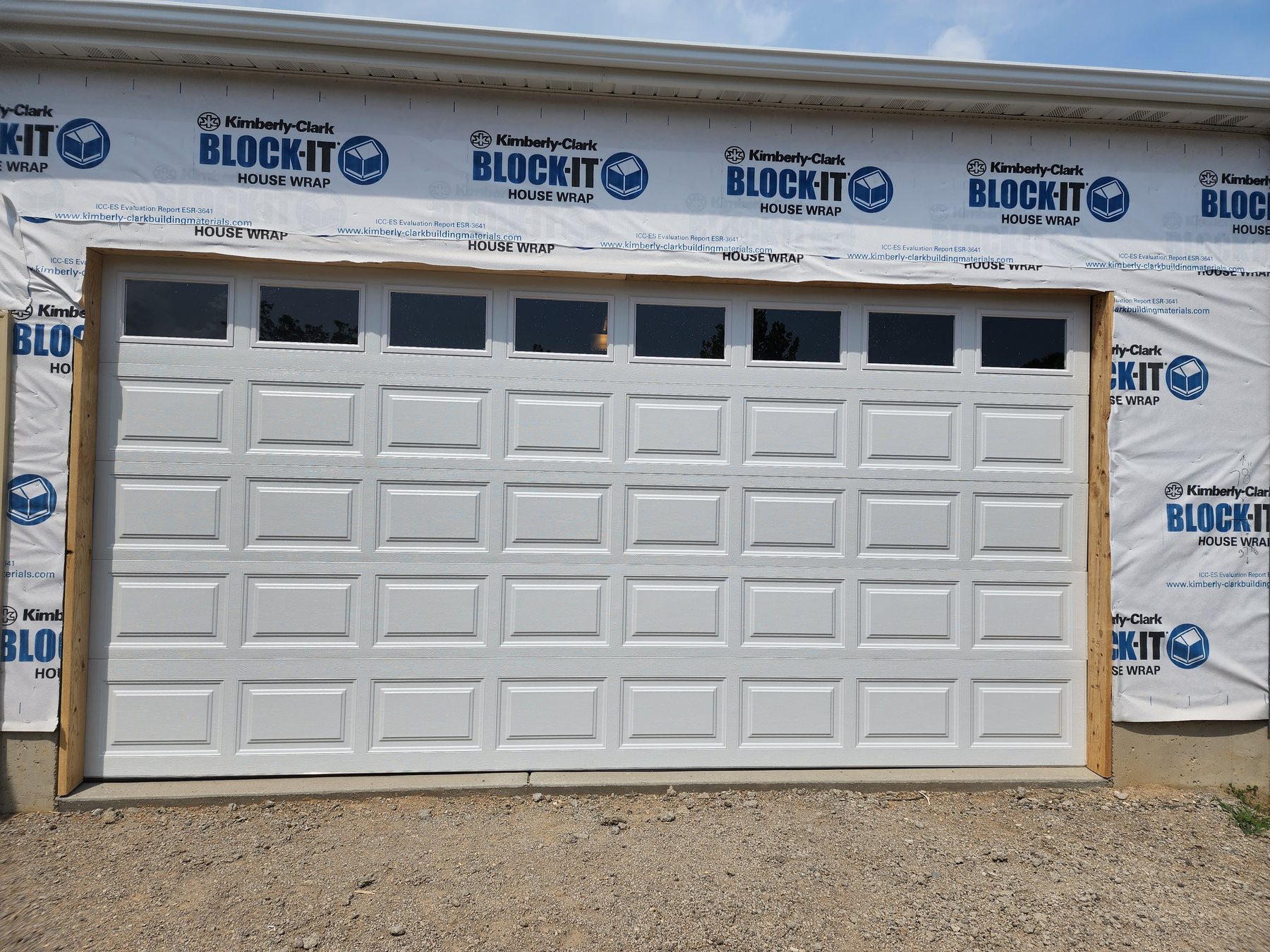 A new white raised-panel garage door with a row of windows on a house still wrapped in house wrap